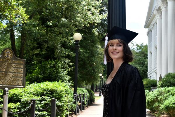 Leah stands on a college campus with many surrounding trees. She is in the center of the photo wearing a graduation cap and making eye contact with the camera.