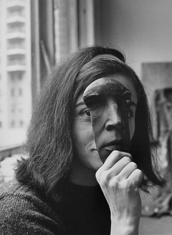 Black and white photo of Marisol Escobar peeking out from behind the sliver of a mask