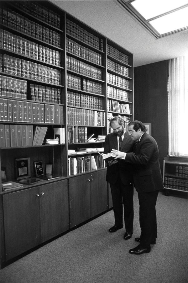 Standing in front of a large bookshelf, Antonin Scalia and C. Ronald Ellington inspect a passage in a book.