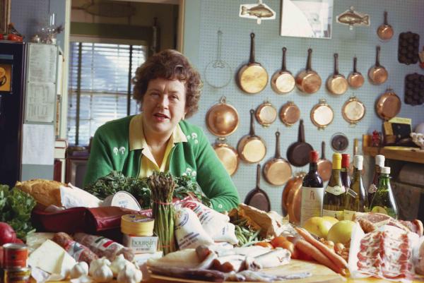Photograph of Julia Child in a kitchen with various foods piled in front of her and a collection of pans hanging on the rear wall.