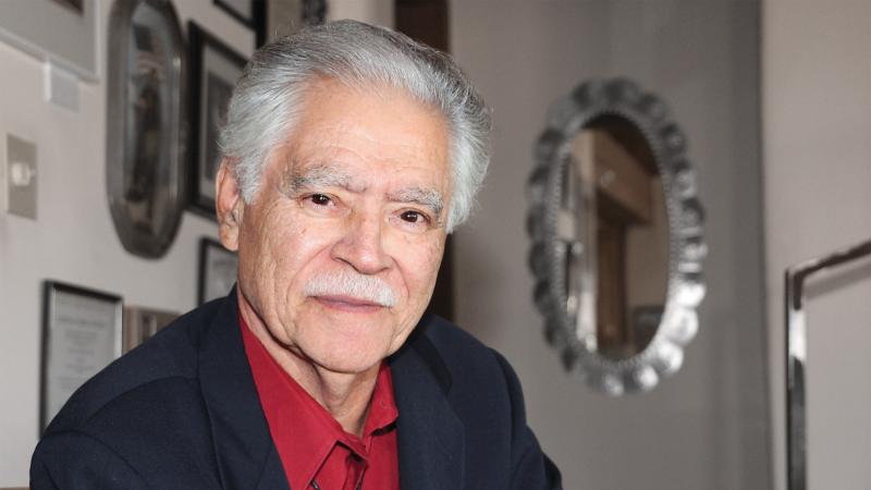 Portrait of Rudolfo Anaya, wearing a red shirt, in his home