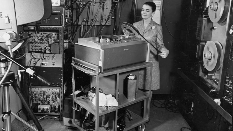 Black and white photo of a woman standing in a room full of machines holding a large roll of film.