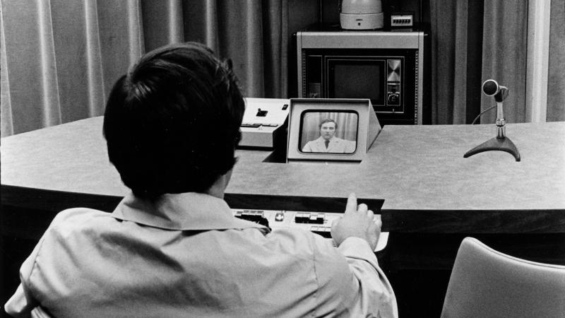 Photo of a man sitting at a desk looking at a small television screen, with a camera filming him in the background.