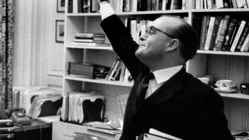 A black and white photo of Truman Capote waving his hand in the air in front of a bookshelf.