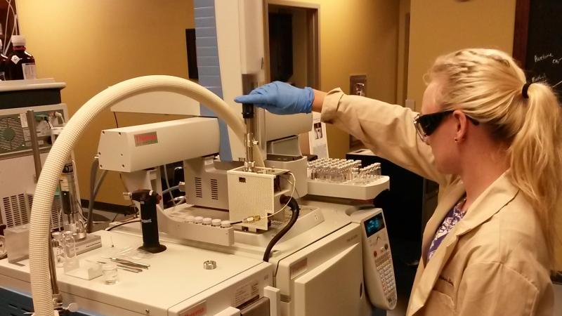 Woman stands at machine in lab coat and goggles