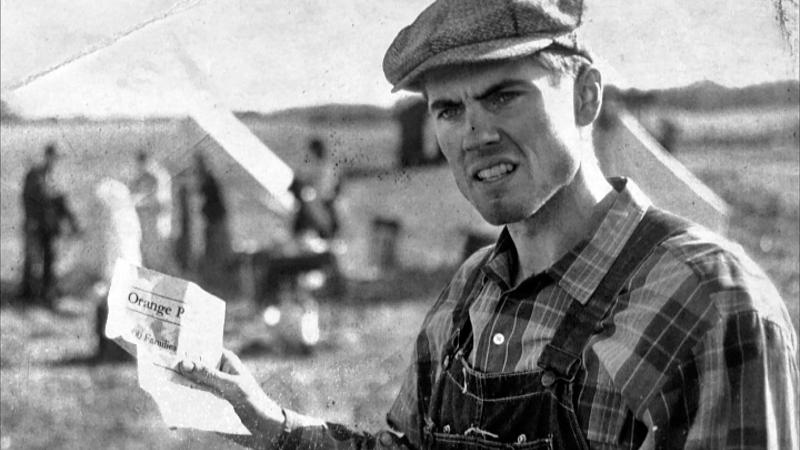 black and white film still of a man holding up a paper, wearing overalls and a hat