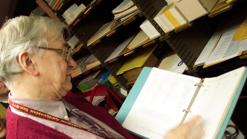 Lampi, wearing a red sweater, reads and holds an open binder in the library stacks