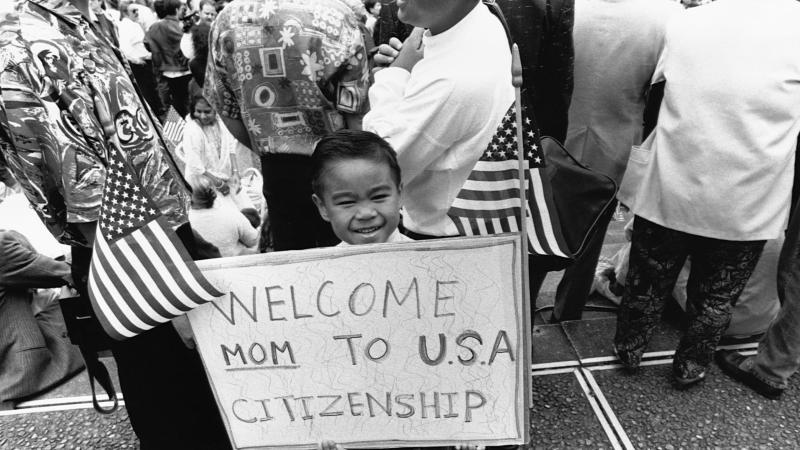 A young boy holds up a sign, flanked by American flags, that reads "Welcome Mom to USA citizenship"