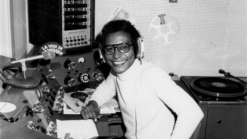 Collinet in a white turtleneck, sitting at a radio broadcasting panel with headphones on, smiling at the camera