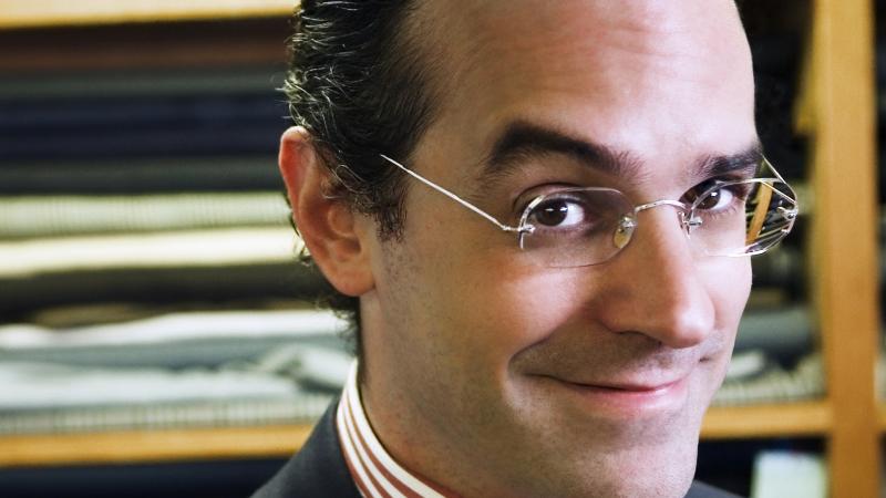 Anton, wearing rimless glasses and hair slicked back, adjusts his blue tie, worn with a red and white striped shirt and black blazer