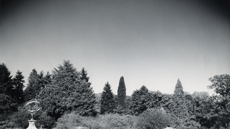 black and white photo of a large garden surrounding by trees, man sitting on bench