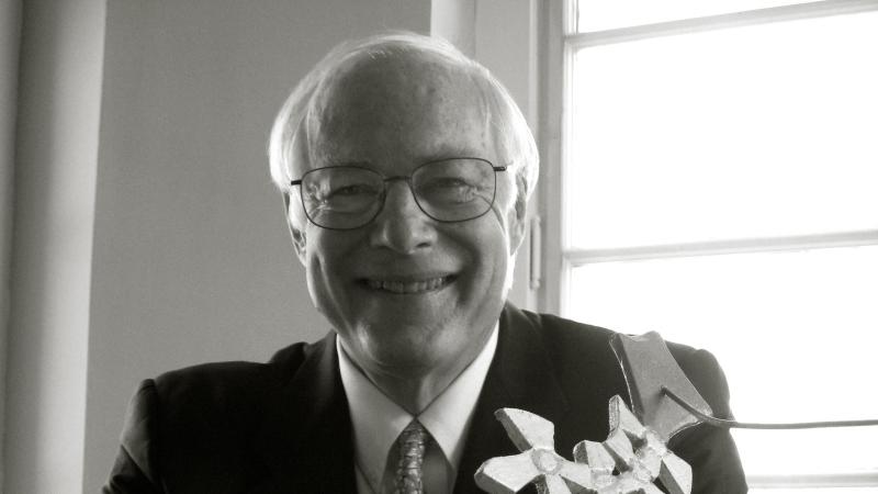 Chairman Leach smiles at the camera, sits with his hands folded on the table in front of him, wearing a dark suit and tie, and eyeglasses 