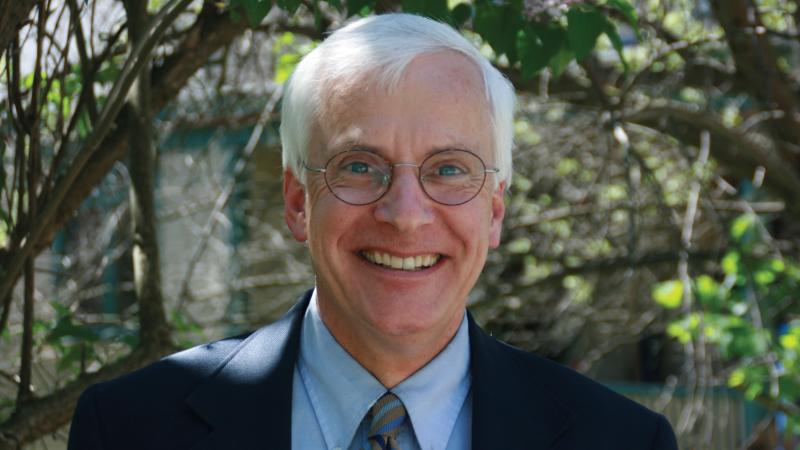 Gilbert smiles at the camera, wears glasses a blue suit and striped tie