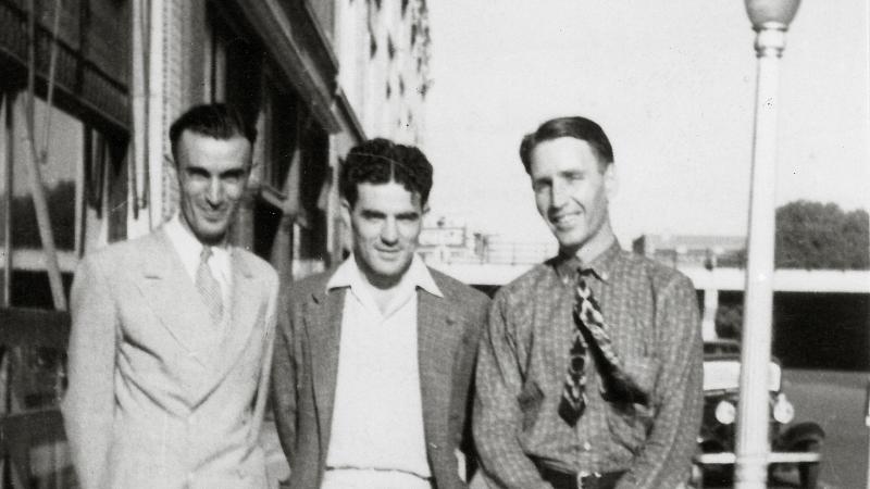 Three young men in light colored suits stand with their arms around each other; Thompson holds a straw hat