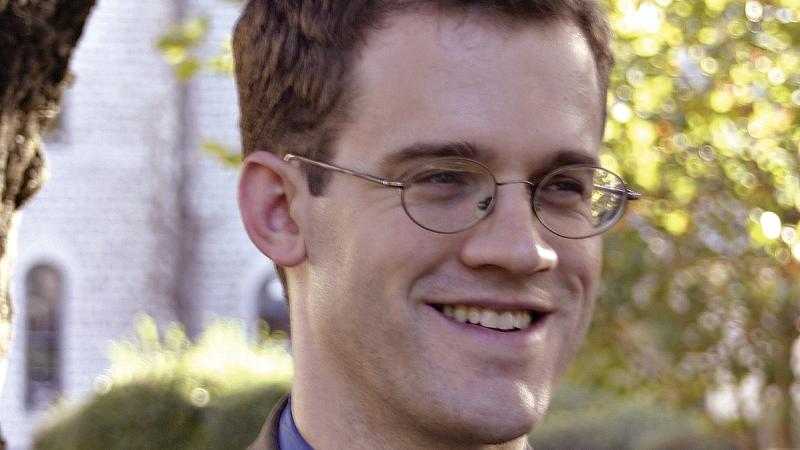 Nel, in a brown suit, blue shirt and eyeglasses, smiles; in the background is a white building and a leafy tree