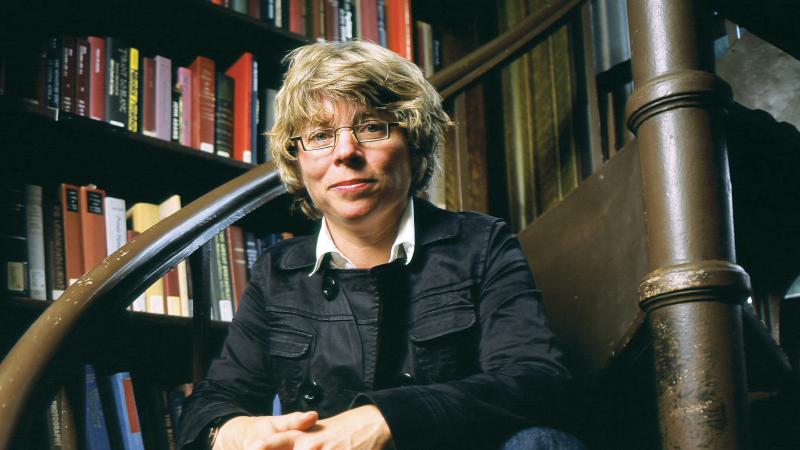 Lepore sits on a step of a winding brown wood staircase, surrounded by shelves of books