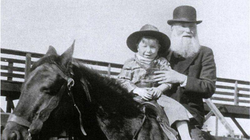 Miller, bearded and wearing a bowler hat, sits behind a young boy on a horse