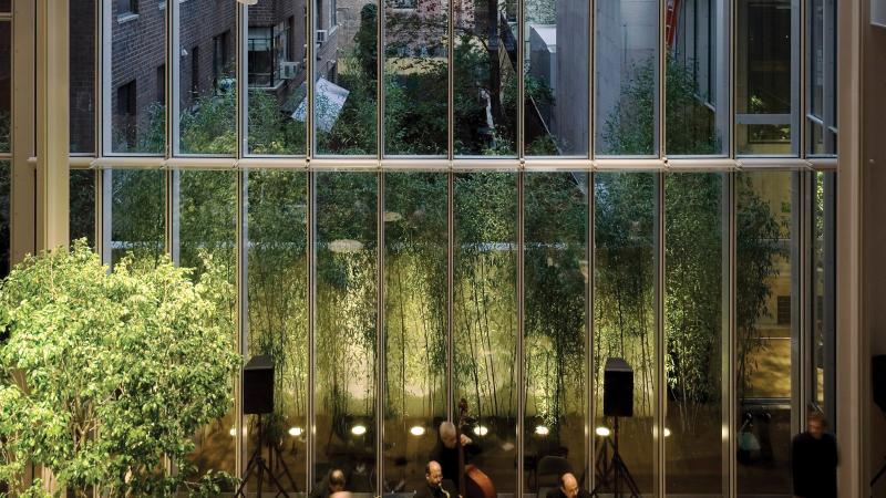 Floor to ceiling windows looking out onto a small green space bordered by tall buildings, a classical quartet plays on a stage in front of the windows