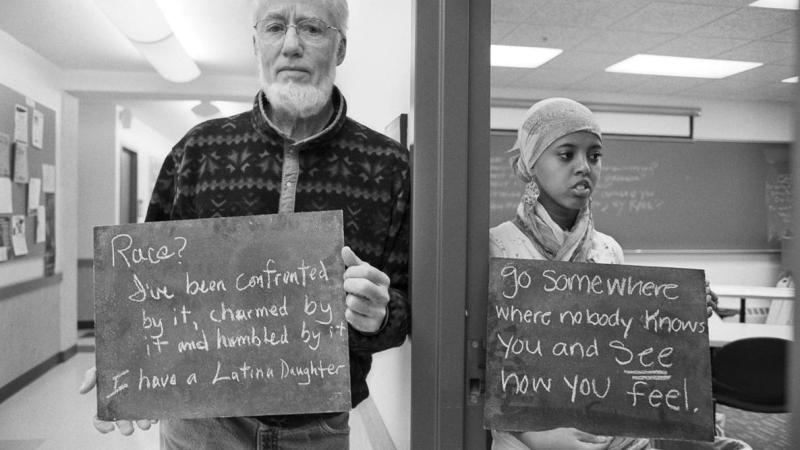 Black and white photo of two individuals holding signs that have provocative statements about race on them.