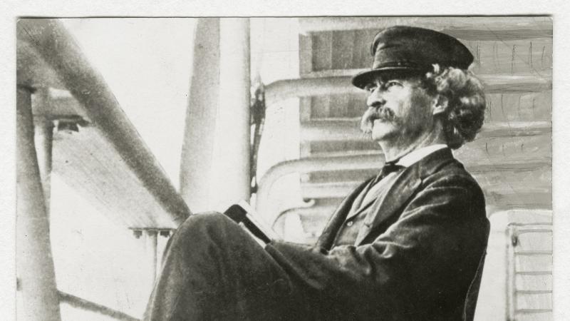Black and white photo of Mark Twain sitting on a chair on the deck of a cruise ship.