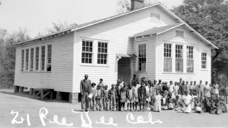Black and white photo of African American students standing in front of a small white schoolhouse.