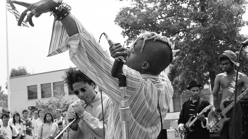 Black and white photo of an African American man performing on stage wearing a striped dress shirt.