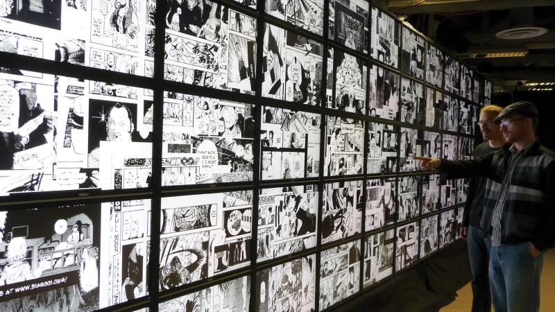 Two men stand in front of numerous small screens lit up with black and white manga illustrations