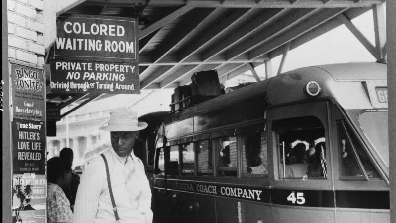 An African American man in a white hat heads toward the segregated waiting room while a bus idles at the curb