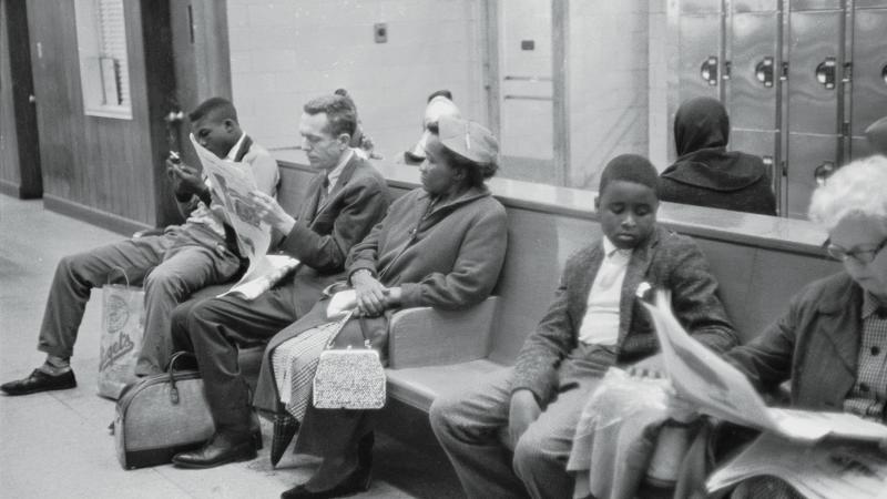 Black and white people sit on a bench next to each other while waiting for their bus