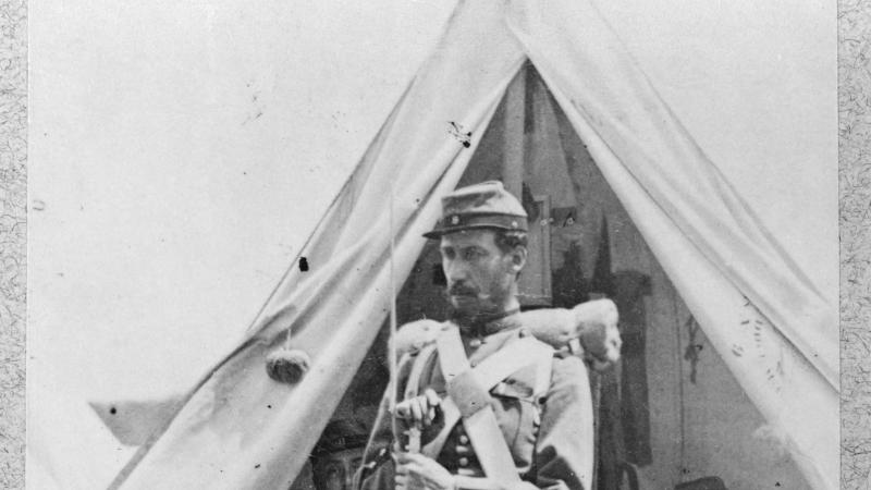 A soldier in full uniform, holding a bayonet, stands in front of a tent, where his fellow soldiers are sitting