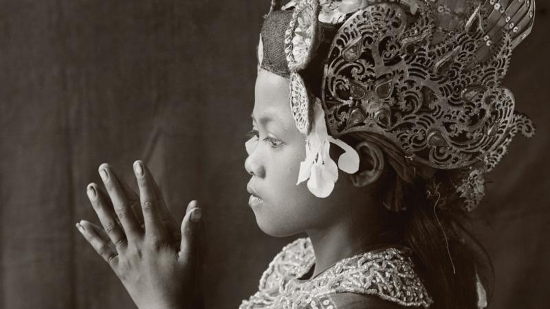 Side view of a young balinese dancer, palms clasped together, wearing a tall floral headdress and embroidered dress