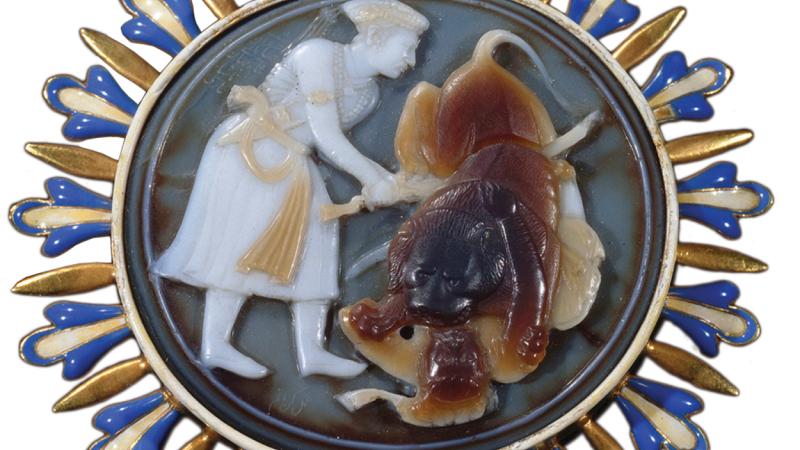 Man made of white ivory being attacked by a lion, done in brown stone. The cameo is set in gold, studded with blue and yellow stones