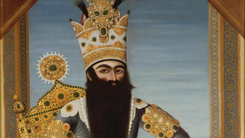 The Shah, with a long black beard, sitting on a richly decorated gold throne, wearing blue robes, gold jewelry, and a tall gold crown encrusted with gems