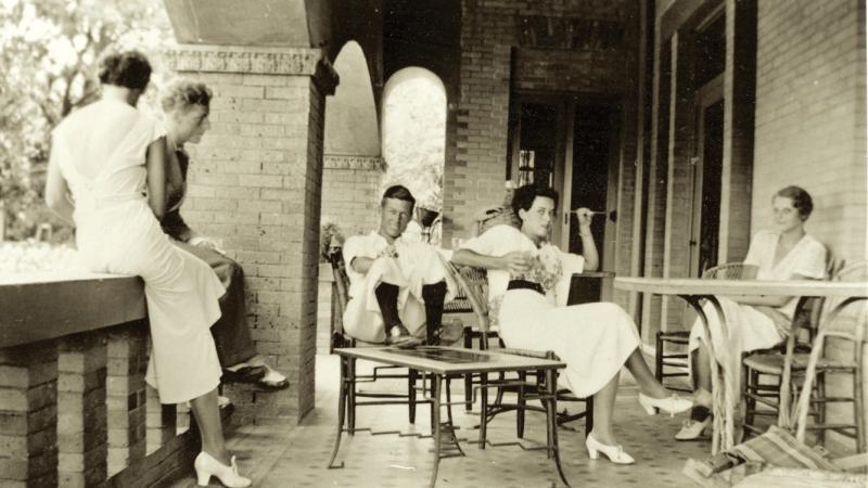 Reed, dressed in white, talking with her friends on the house terrace, sitting in lawn chairs