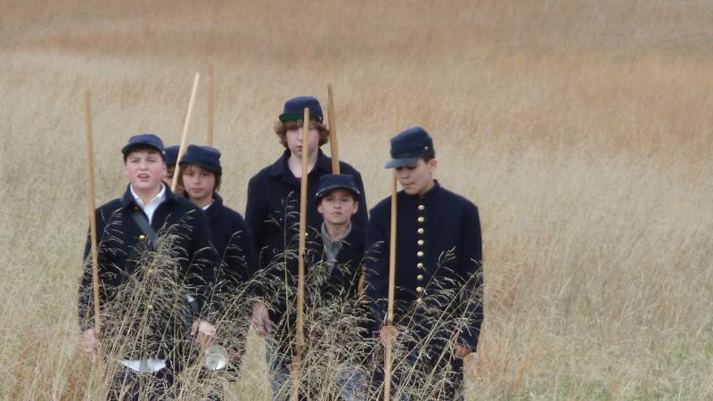 Color photo of a group of schoolboys re-enacting a Civil War march through a field. 