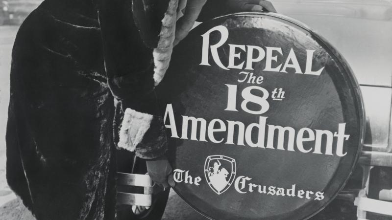 Black and white photo of a woman standing before a spare tire cover, on the back of a car, that says, "Repeal the 18th Amendment."