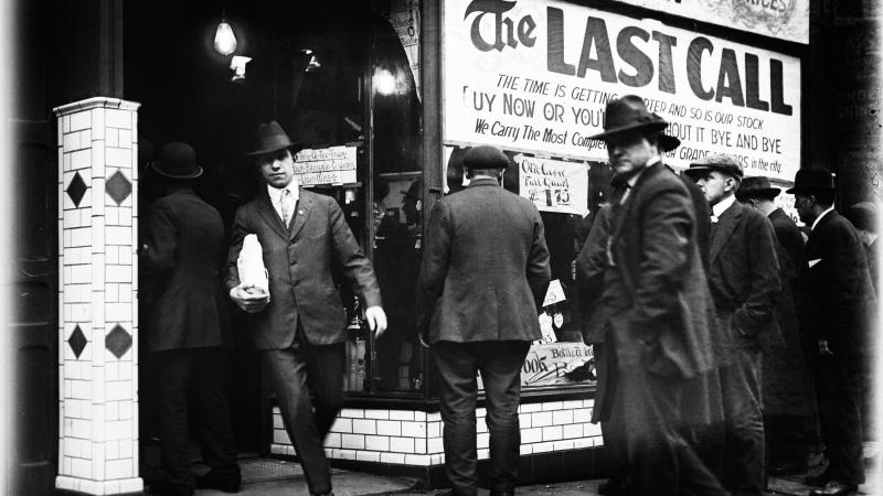 Black and white photo of men in formal attire heading into a liquor store. One man carries a packaged bottle in the foreground. 