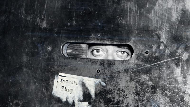 Black and white photo of a man peering through a door slat, as if he were reconnoitering the area.