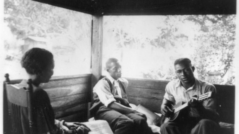 Black and white photo of three African Americans sitting on a porch, one of them playing the guitar.