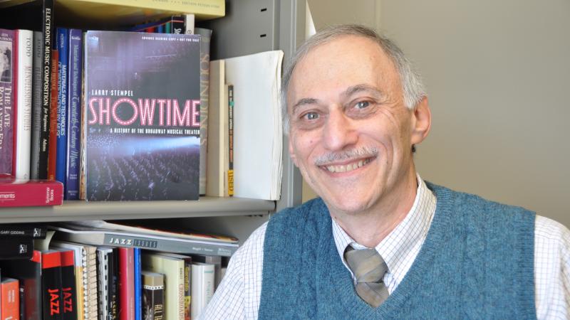 Color photo of Larry Stempel next to a bookcase, wearing a dark green vest, shirt, and tie.