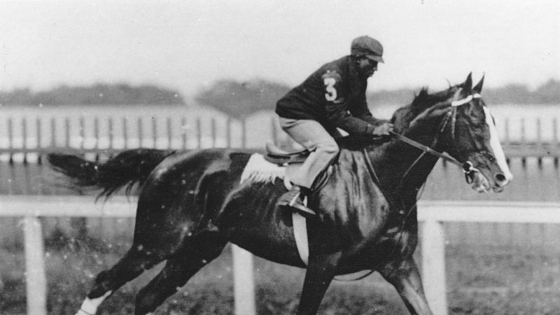 Black and white photo of an African-American jockey riding a horse at high speed. 