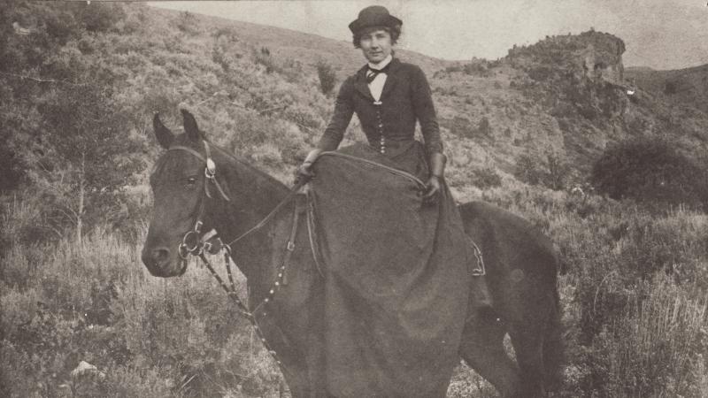 Black and white photo of a woman riding a black horse, wearing a full dress and hat.