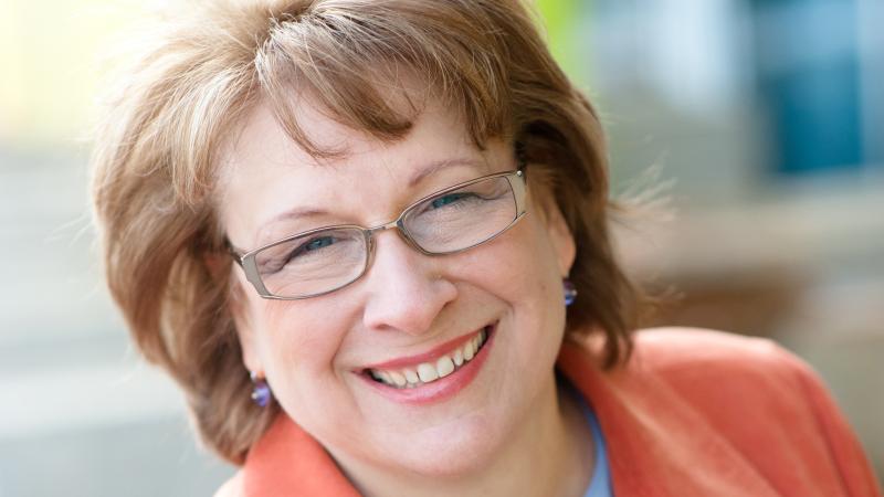 Color photo portrait of Maggie Coval in a long-sleeved orange blazer.