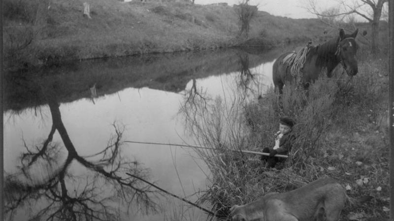 Black and white photo of a little boy fishing in a stream while his dog attempts the same maneuver in the foreground.