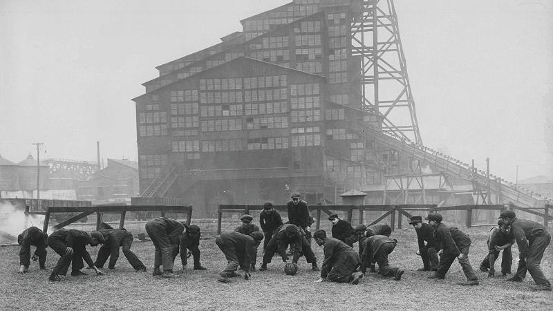 Black and white photo of boys playing football with a mining facility in the background.