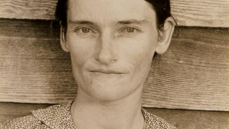 Sepia-colored photo portrait of a sharecropper woman against the wood siding of a house.