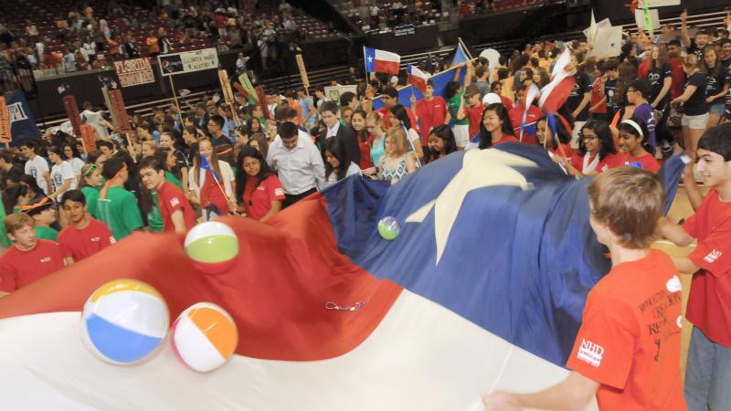Color photo of a crowd of young students flopping inflatable balls around on top of a large Texas flag.