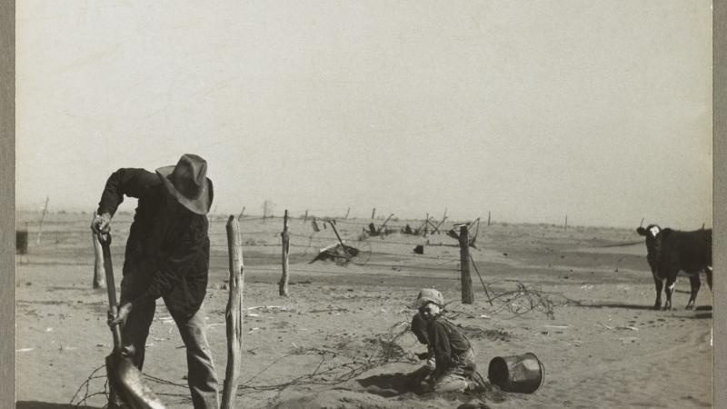 Black and white photo of a man shoveling sand in the foreground, with a child to his right, sitting on the plain, and a cow further in the distance.