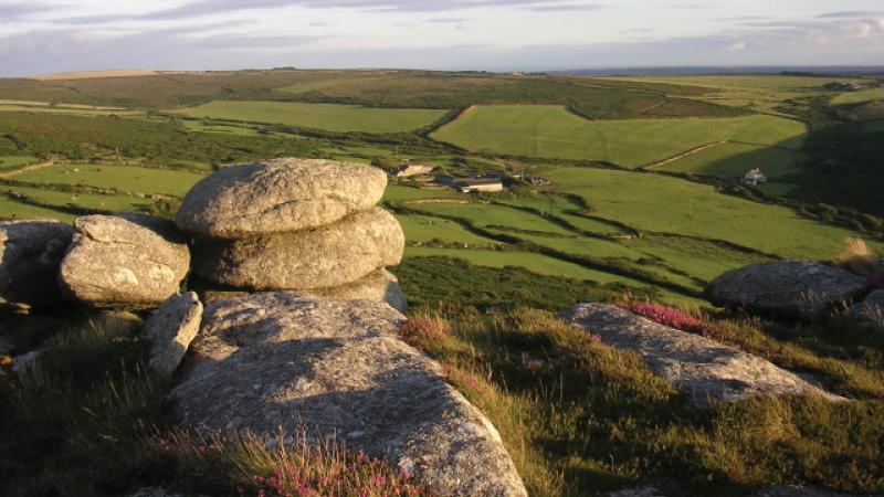 Lush green farmland extends into the distance below a hill covered in stones and grass, with late afternoon light