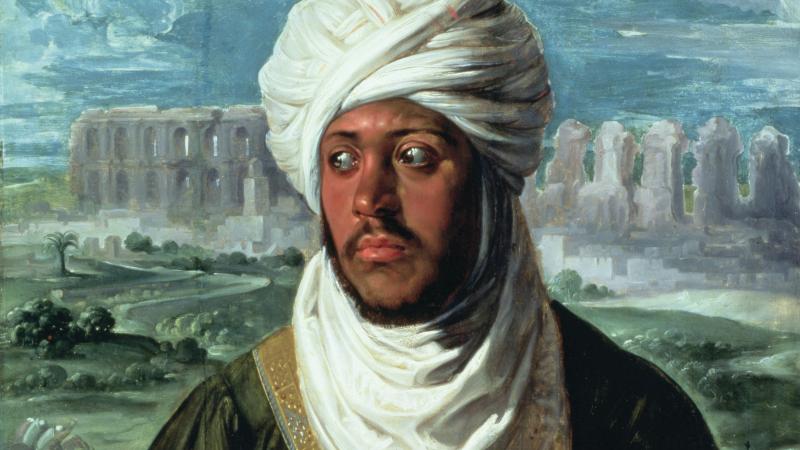 Ahmad in a white turban, green robes, standing in front of green pastures on white stone buildings